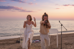 Two people perform with microphones on a beach at sunset, seated on white chairs with the sea and sky in the background. A third microphone stand is set up nearby. • Eleonora Fisco