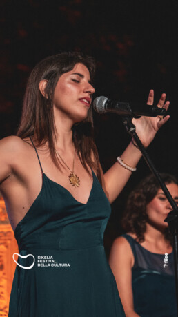 A woman in a teal dress sings into a microphone with her eyes closed and arms raised. Another woman stands in the background. Festival logos are visible on the image. • Eleonora Fisco