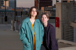 Two people stand close together on a rooftop parking lot; one wears a teal coat and the other a dark jacket, with urban buildings in the background. • Eleonora Fisco