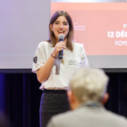 Una giovane donna è in piedi con un microfono in mano, mentre parla di fronte a un pubblico con uno schermo di presentazione proiettato dietro di lei. • Eleonora Fisco