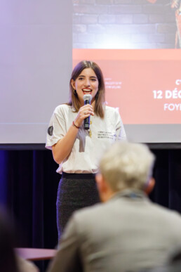 Una giovane donna è in piedi con un microfono in mano, mentre parla di fronte a un pubblico con uno schermo di presentazione proiettato dietro di lei. • Eleonora Fisco