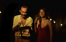 A man and a woman stand side by side holding microphones, smiling, and appearing to perform or speak on stage under warm, dim lighting at the Poetry Slam a Pisa. • Eleonora Fisco