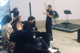 A person stands and reads from a book to a seated audience in a gallery-like space during the Poetry Slam a Pisa, while another person films the event. Audio equipment is set up on a small stage nearby. • Eleonora Fisco